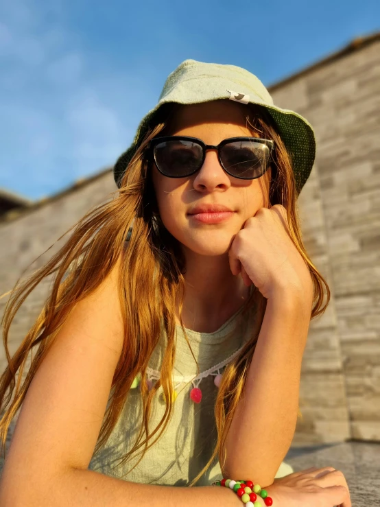 young woman sitting on brick wall wearing sunglasses and a hat