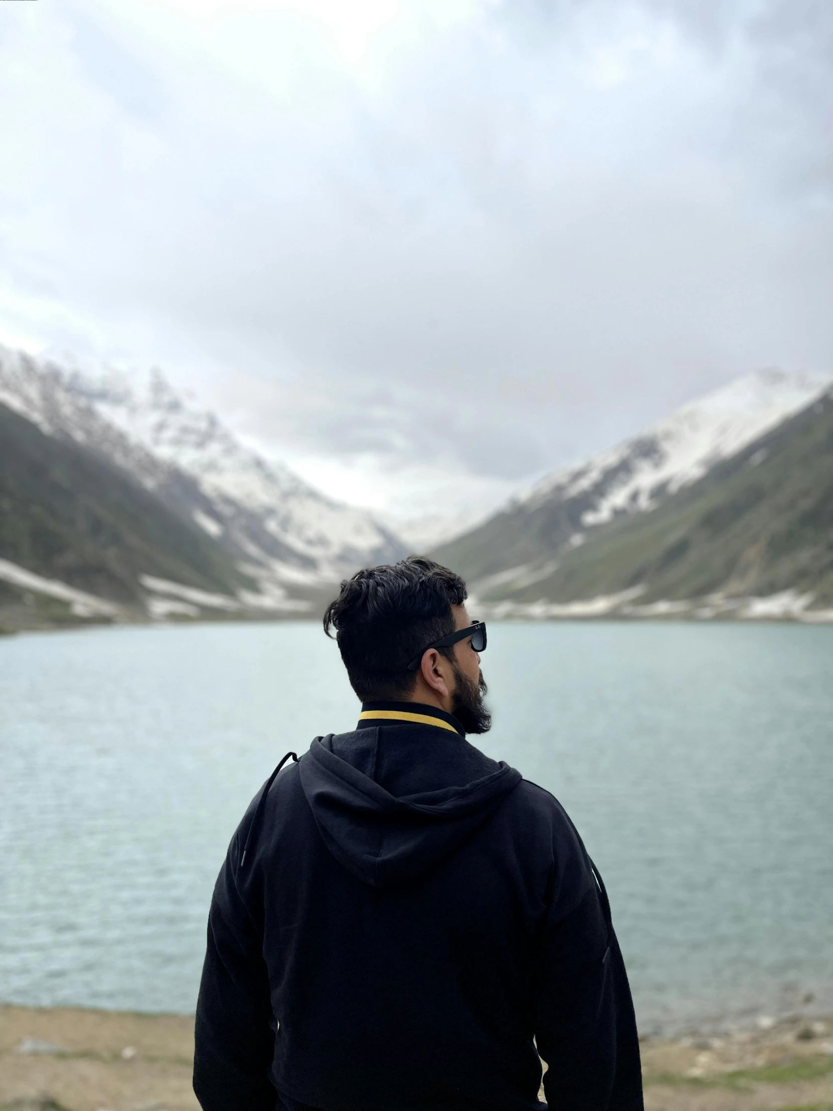 a man with a beard stands looking out at mountains and the water