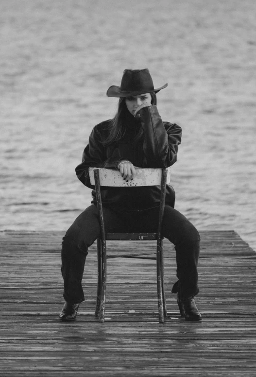 a man in a cowboy hat and black jacket sits on a chair at a dock