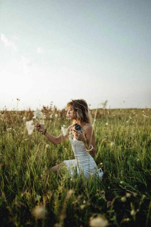 a young woman kneeling in the middle of a field blowing on dandelion