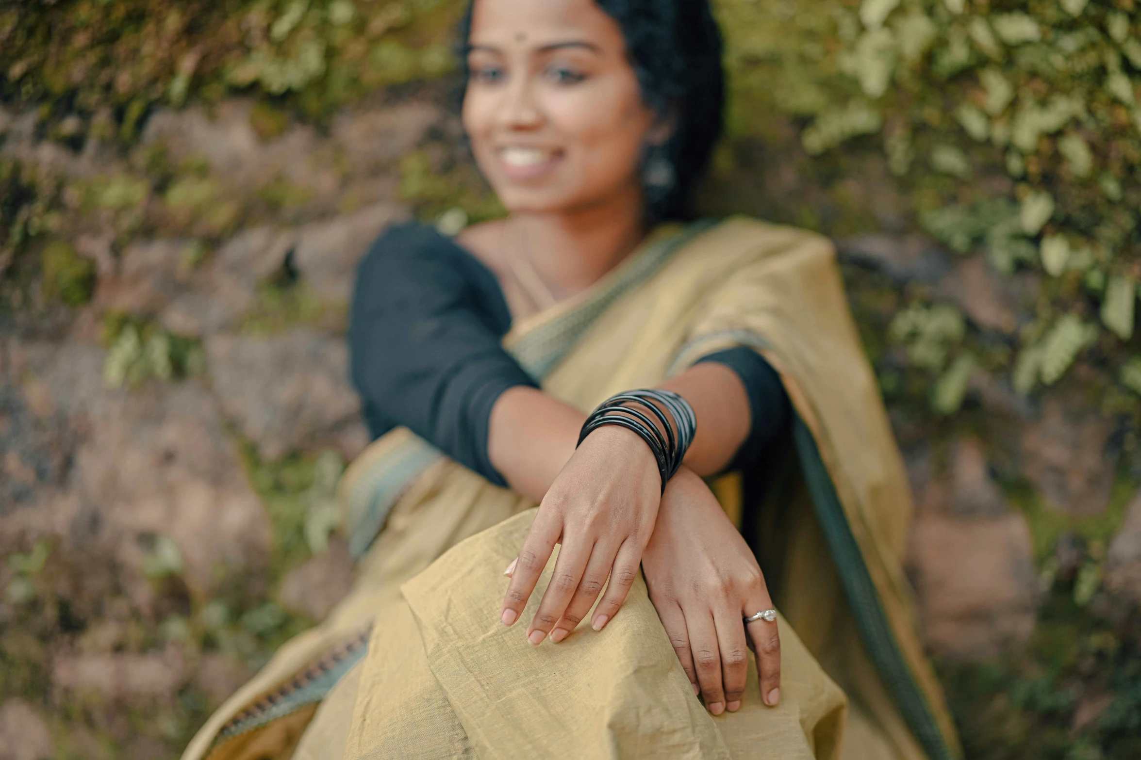 a smiling woman in a black dress and celets poses with her hands wrapped around the back of her purse