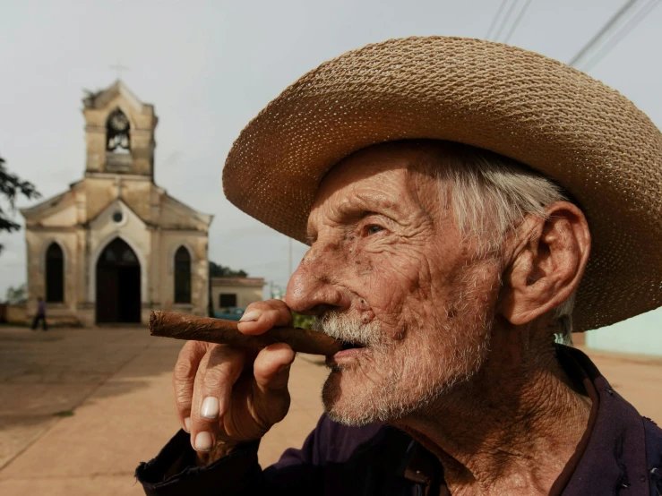 an older man with grey hair and wearing a hat and holds a cigar in his mouth while standing outside a church