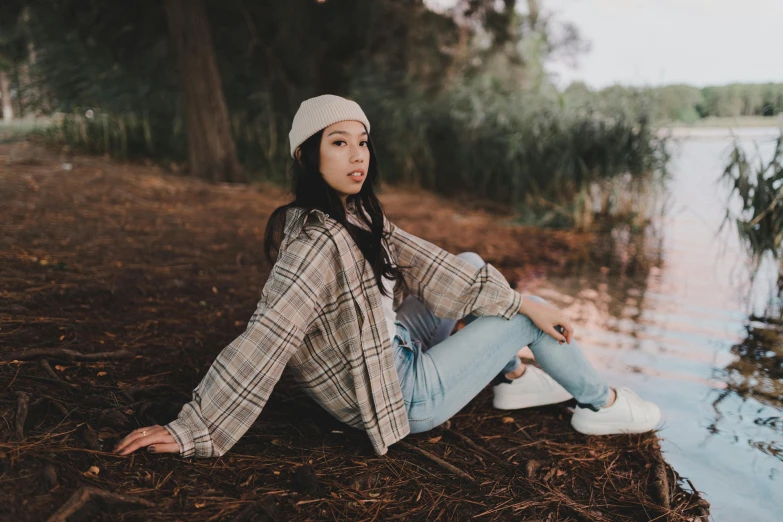 a girl sits by the water wearing jeans and a hat