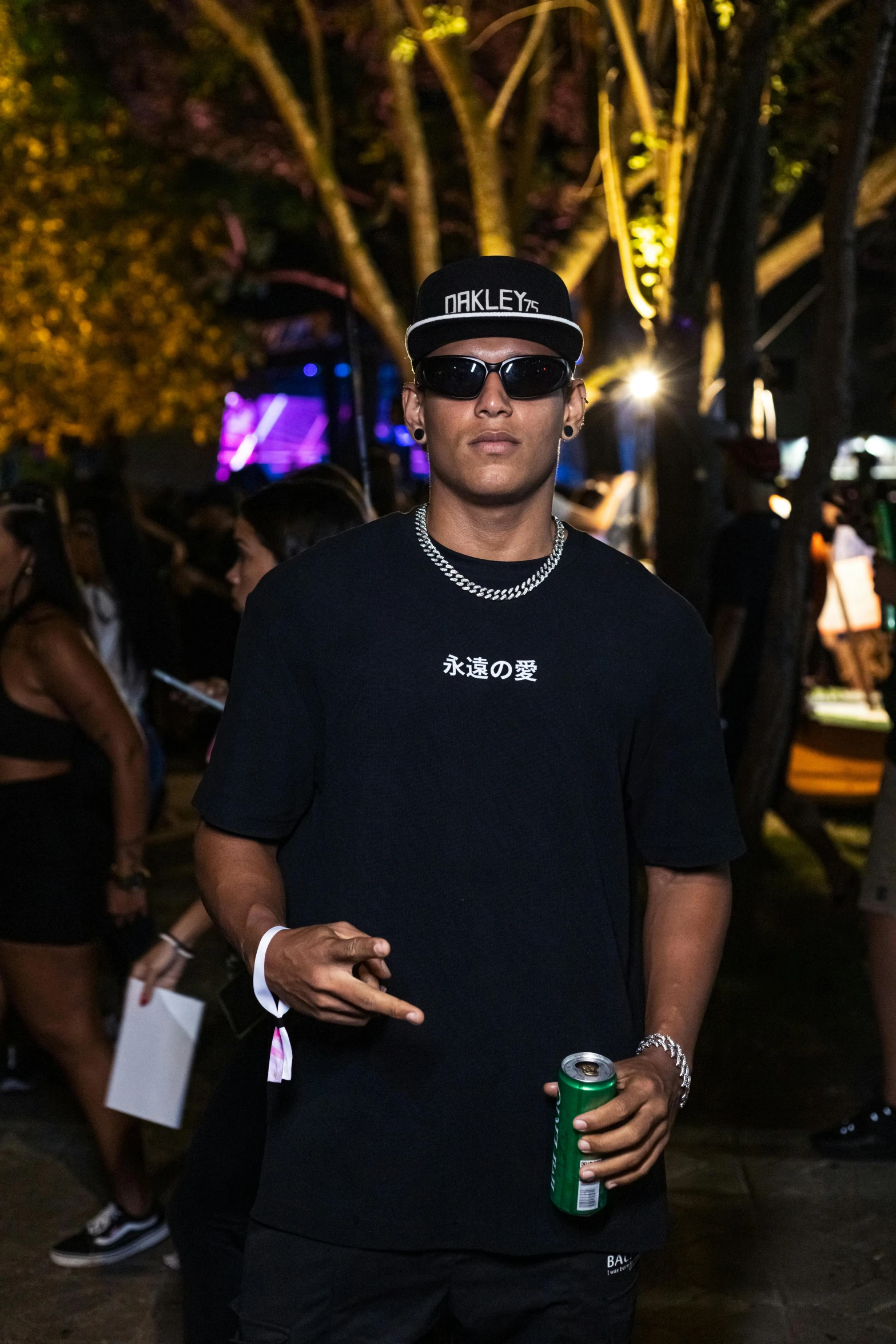 young man in black shirt with baseball cap and sunglasses standing near a group of people at night