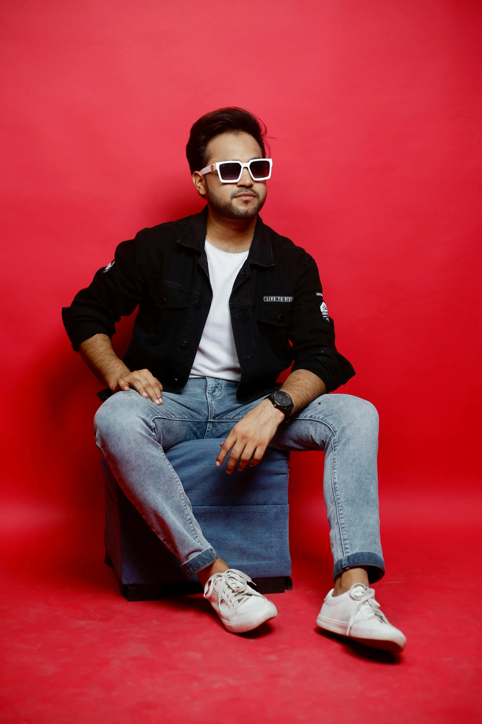 man in denim sitting against a red backdrop with a pair of sunglasses