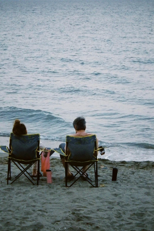 two people are sitting on folding chairs at the beach watching a wave crash into shore