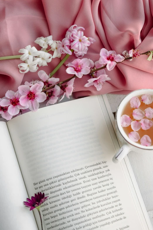 a cup of tea sits next to an open book with flowers and a pink cloth