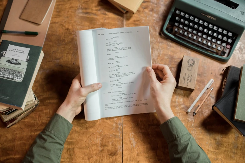 person is reading an open book with some typewriters and pens