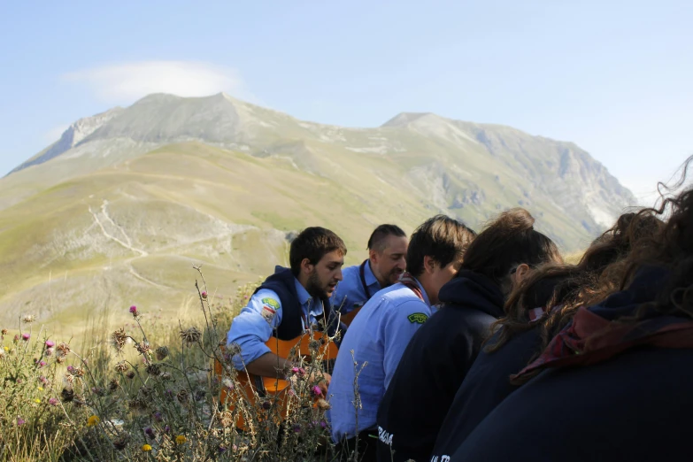 two men are examining a bunch of wildflowers with a mountain in the background