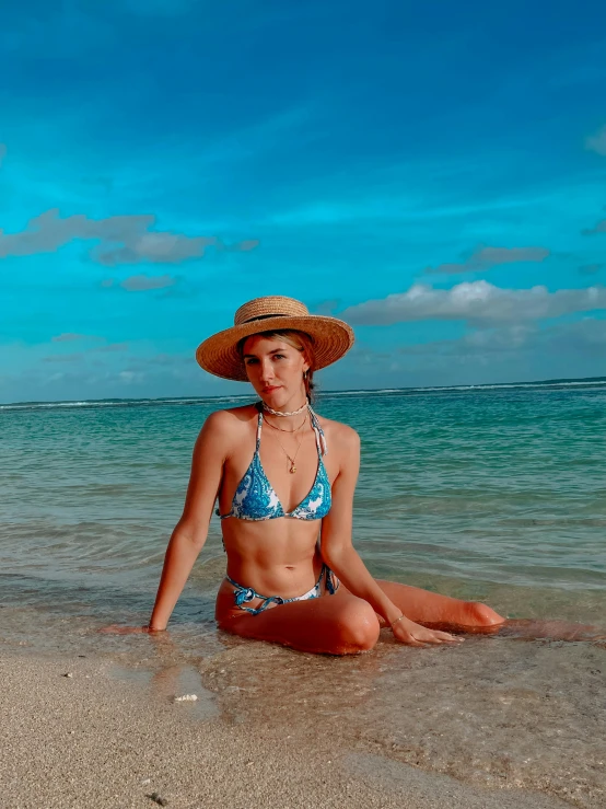 a beautiful woman in a bikini and straw hat sitting on the sand