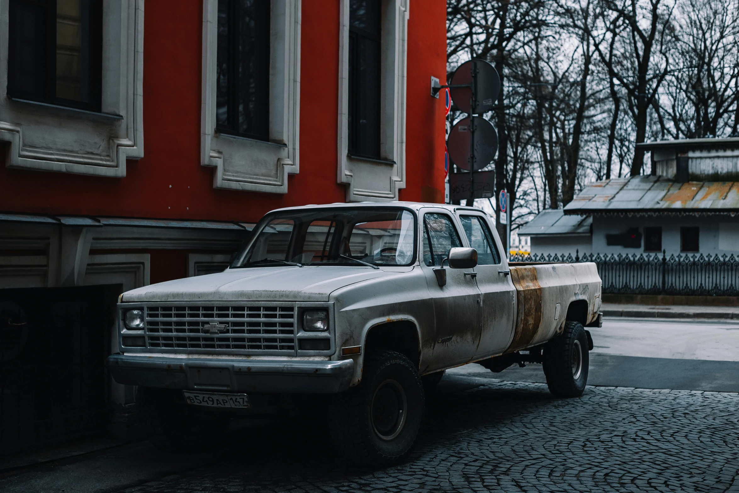 an old white pickup truck with rusted rims sits near a red building in the city