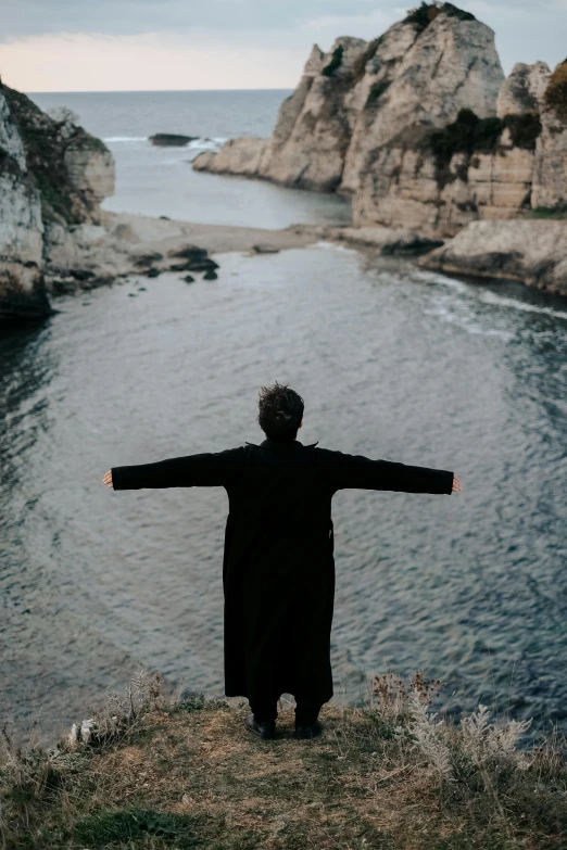 a man in black standing on top of a cliff by the ocean