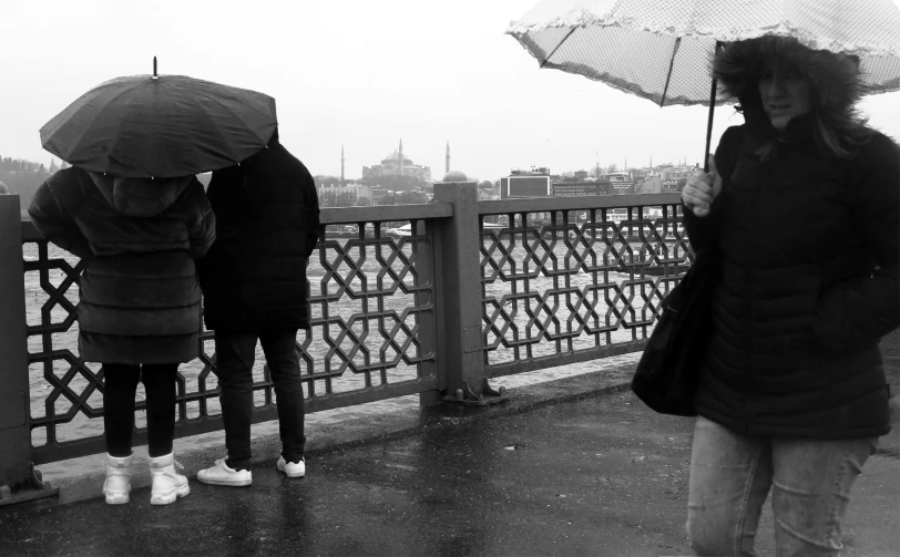 three people with umbrellas stand in the rain next to a fence