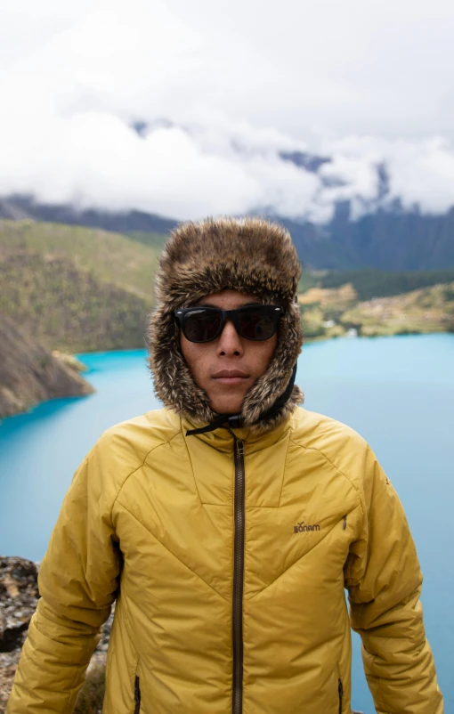 a man in sunglasses and a coat stands by a mountain lake