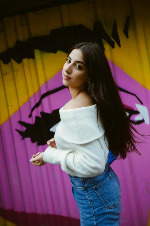 woman with long dark hair and blue jeans leaning against a colorful painted wall