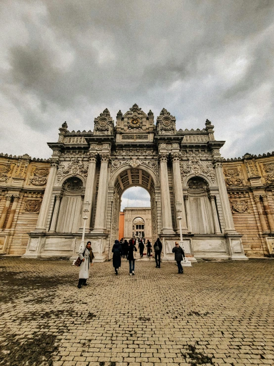 people walk past a large stone arch on a cloudy day