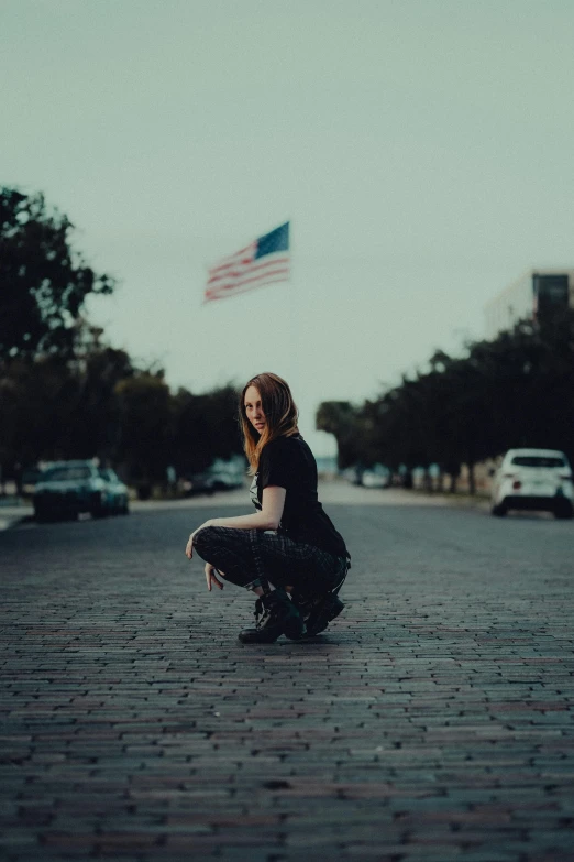 a woman crouches down on the side of a road, with an american flag in the background