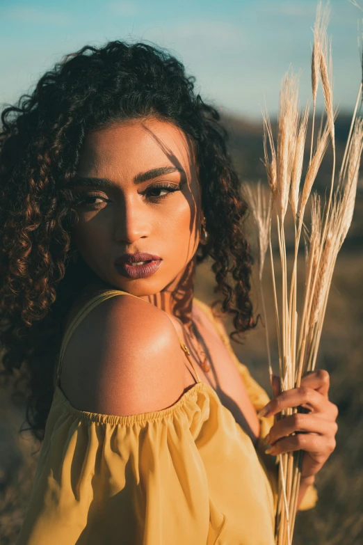 a close - up of a woman in a yellow dress with a hair and make up holds wheat stalks