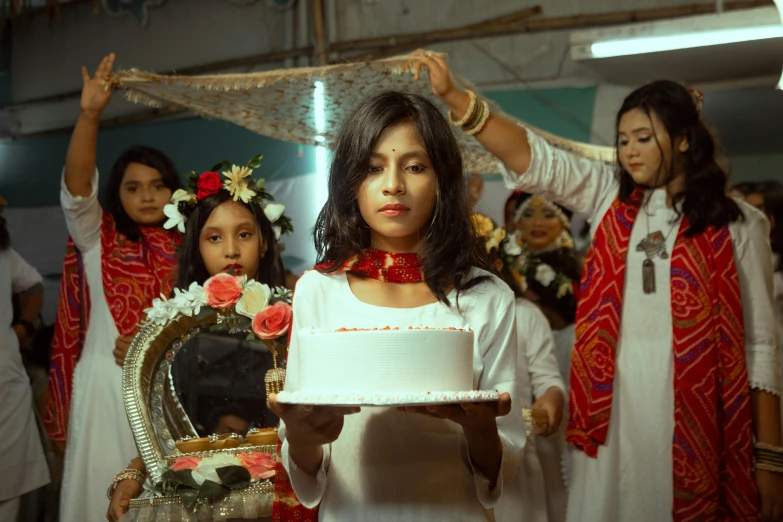 people in white dresses and gold headdresses hold a white cake with red flowers on it while others in costume stand behind her