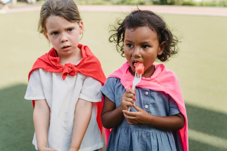 two children are outside in their costumes, one with food on his tongue