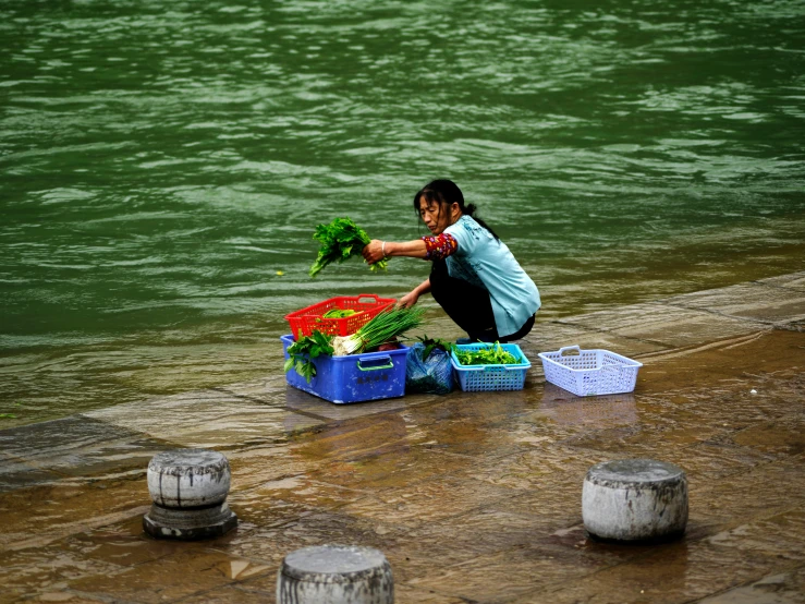 a woman is squatting down with vegetables in containers next to the water