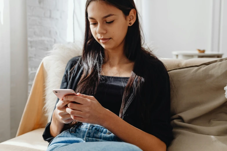 a girl in a black shirt and jean pants sits on a couch holding a smart phone