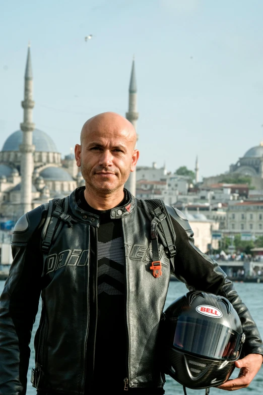 man wearing a helmet and jacket by water with a large city behind