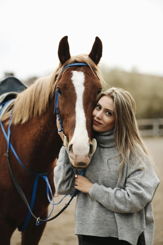 a woman in a gray sweater hugging a brown and white horse