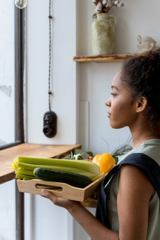 a woman holds a box filled with cucumbers, oranges, and green beans