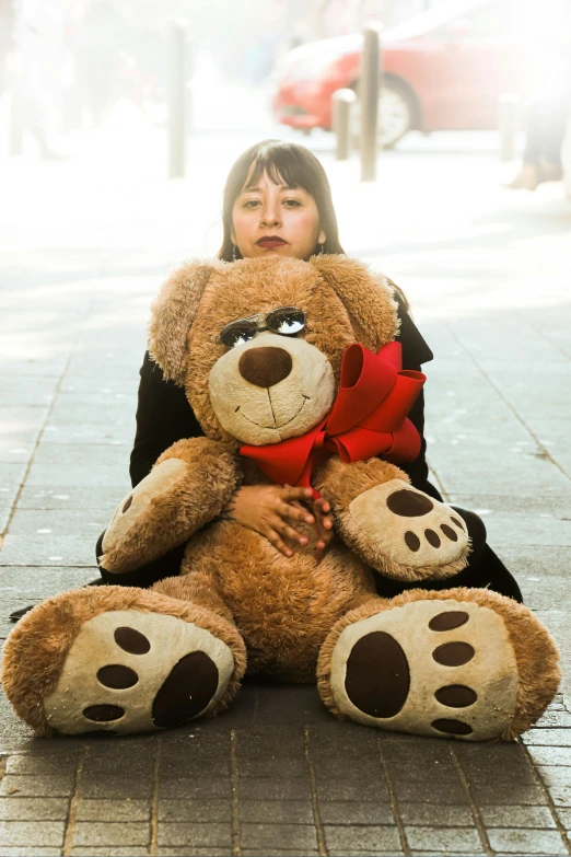 a woman is sitting on a sidewalk holding a large stuffed bear