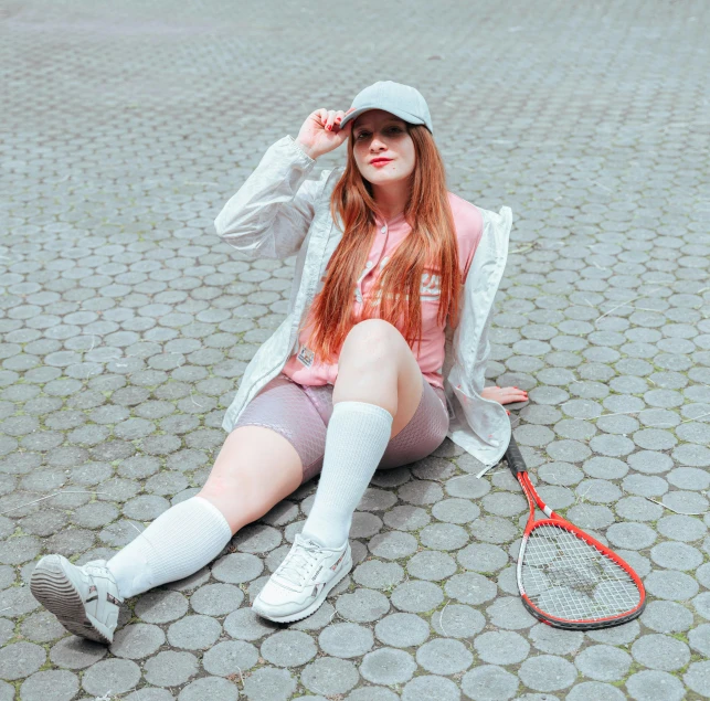 woman in pink sweater, grey shorts, and white socks sitting on ground next to tennis racket