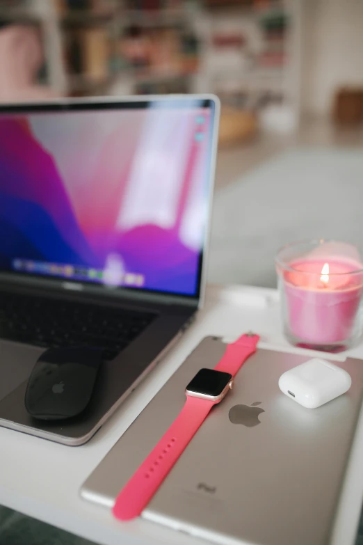 the view of an apple laptop and a pink apple watch on a desk with a candle, tea cup, and a small candle holder