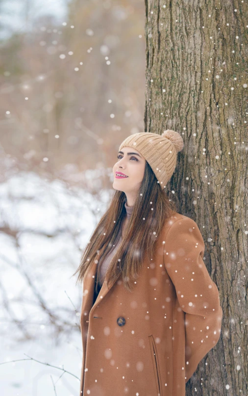 woman leaning on a tree in snow while wearing a hat