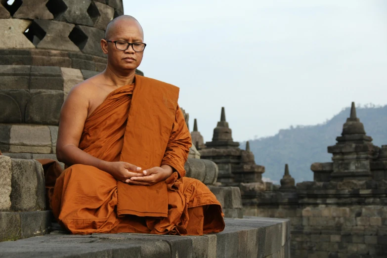 an old man with glasses sits on the edge of a brick pillar in front of old buildings