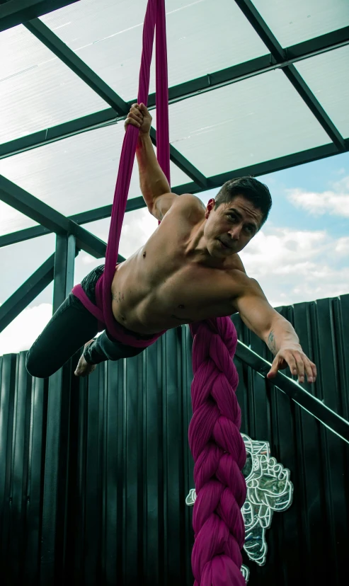 a shirtless man hangs from a rope with a large pink rope in front of him