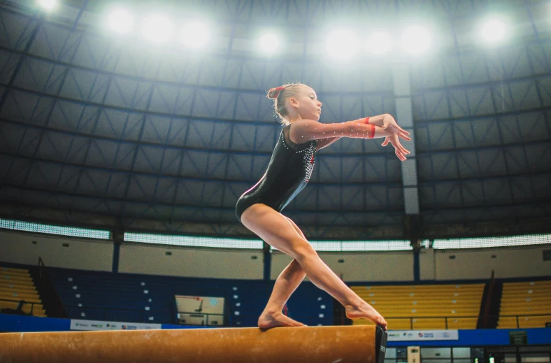 a girl in a black and gold gymnastics outfit, is balancing on a balance beam