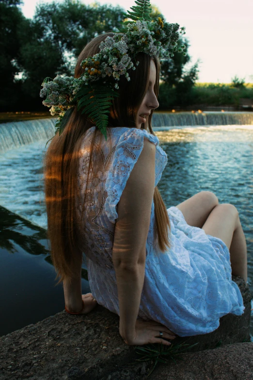 a woman in a flower crown sitting on a rock next to a body of water