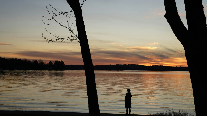 the man is standing by the lake looking at the sunset