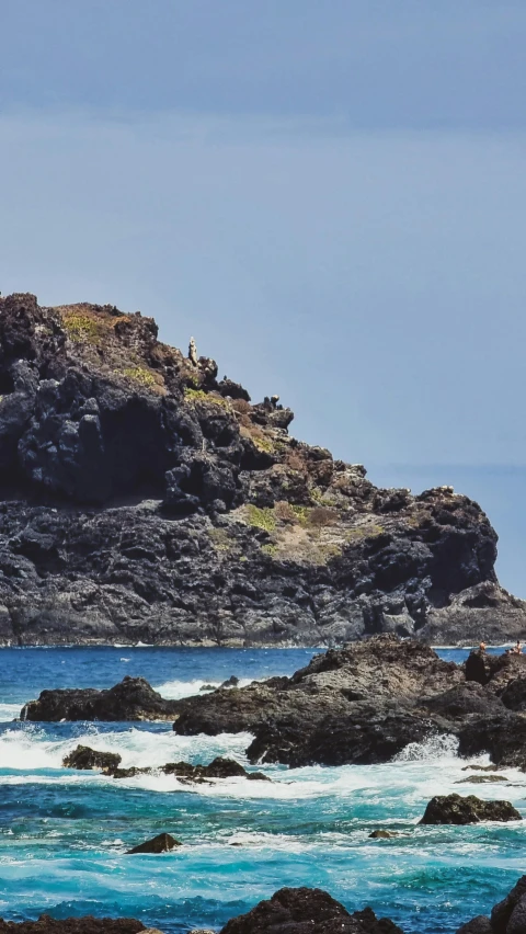 two people stand on rocks near the ocean and look out at the rocky coastline