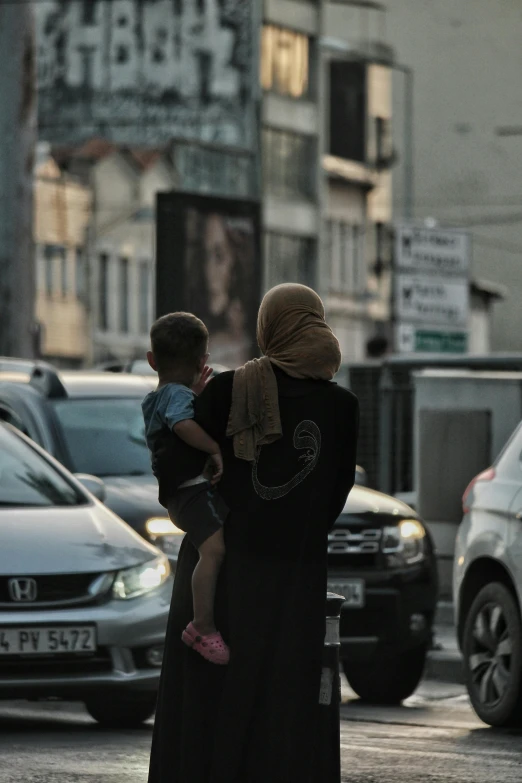 a woman in hijab holds a small boy while looking away from parked cars