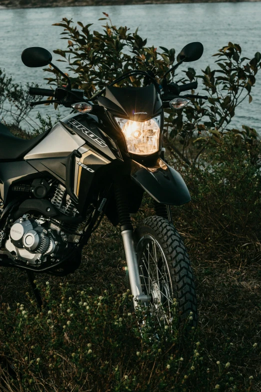 a black motorcycle with the front lights on sits parked on grass next to the water