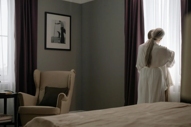 there is a young lady standing by a window in a bedroom