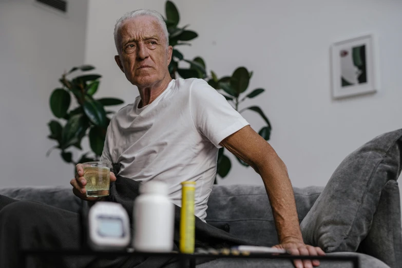 an older man sits on a sofa with his laptop and glass in hand