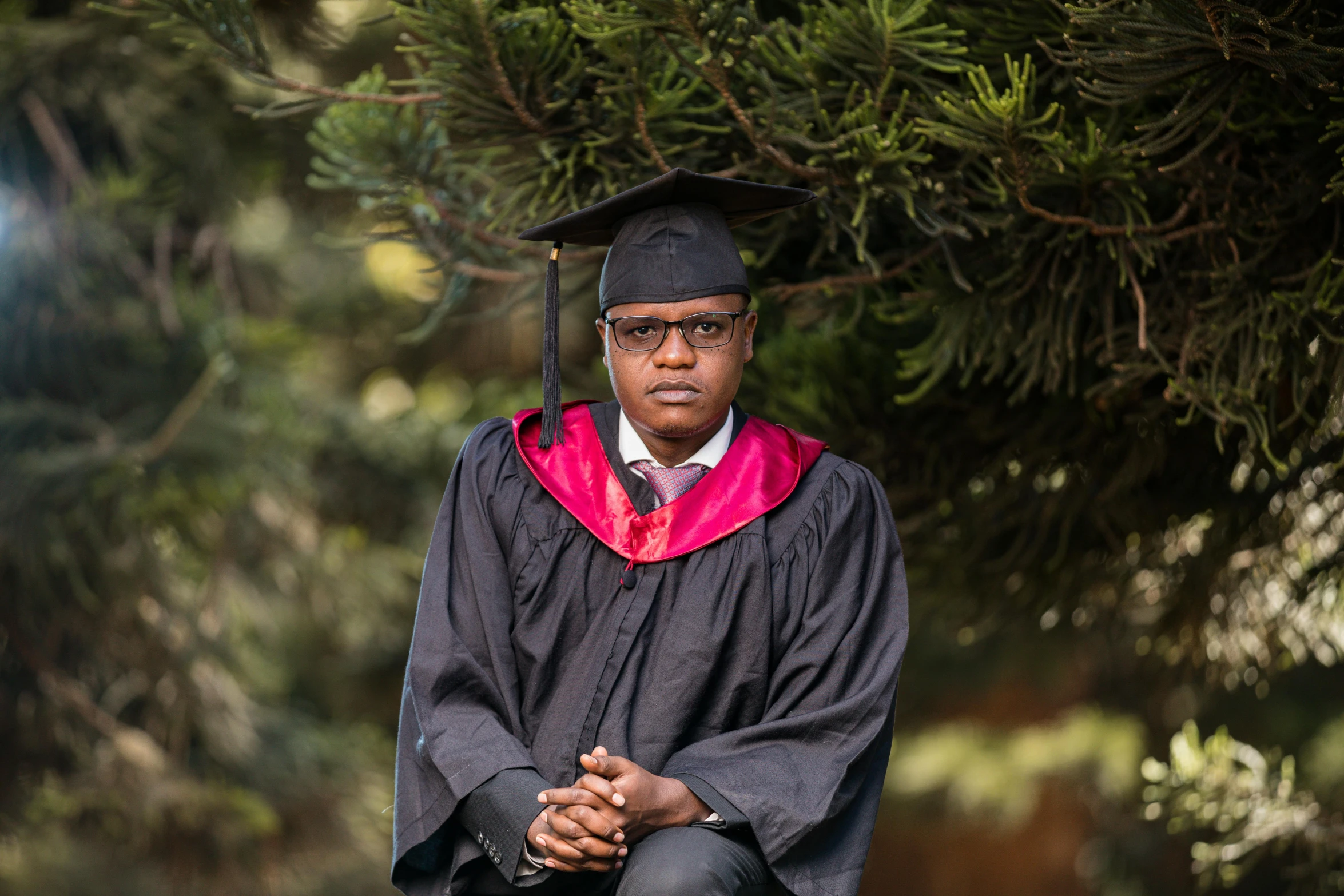 a person wearing a black and red graduation gown and hat sitting with his hands folded