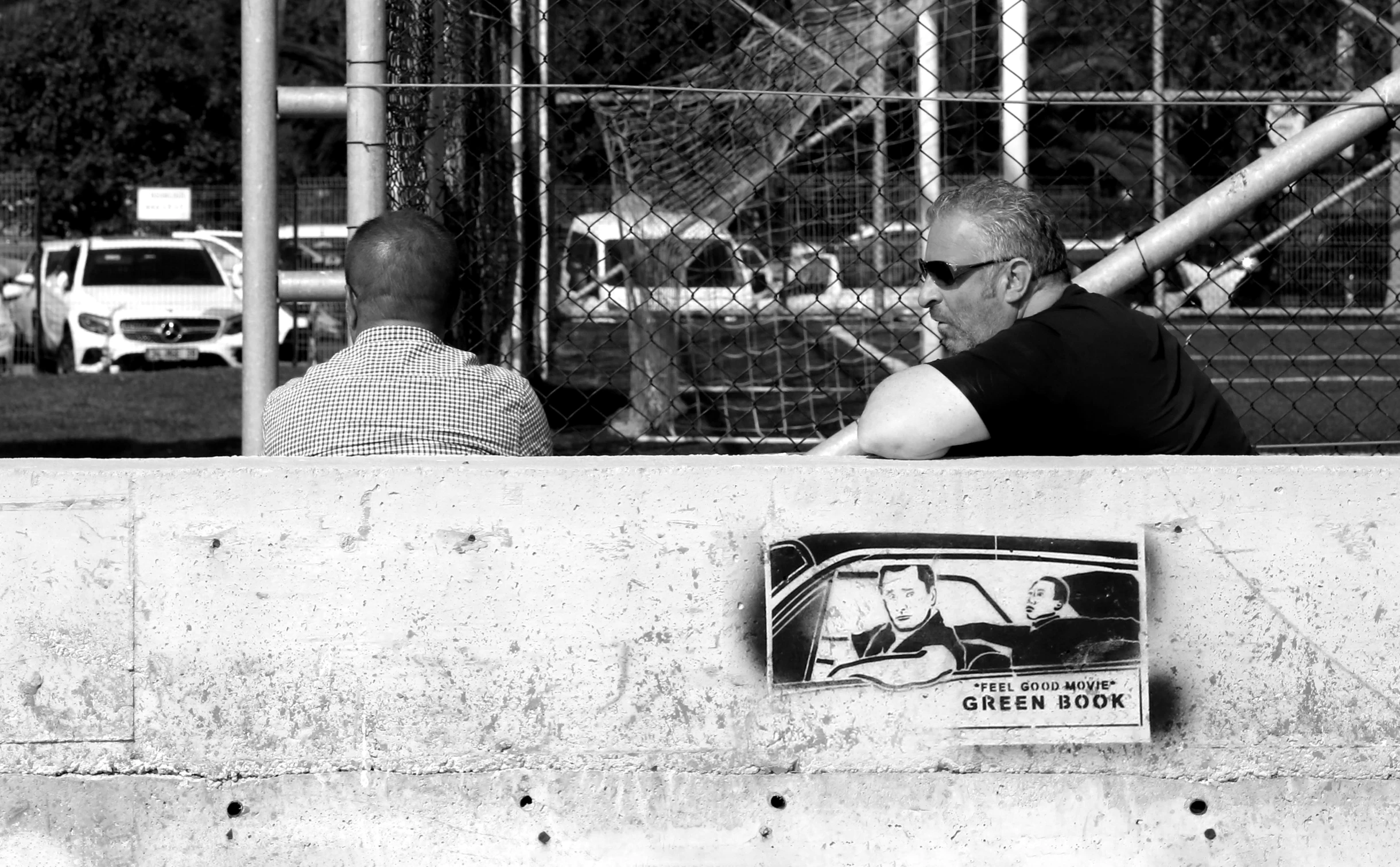 man sitting on fence looking at magazine with a sign and car on it