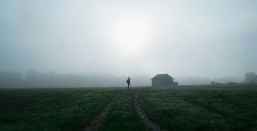 the silhouette of a person standing in front of a misty field with a small house