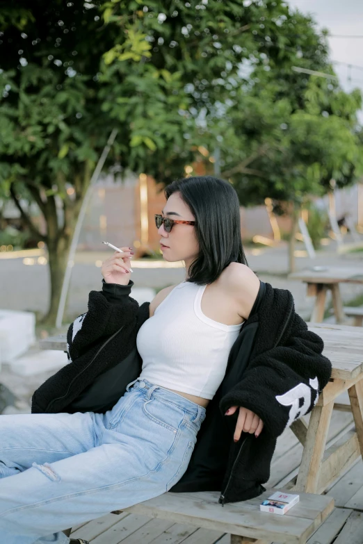 a woman wearing jeans, white top and black jacket smoking a cigarette