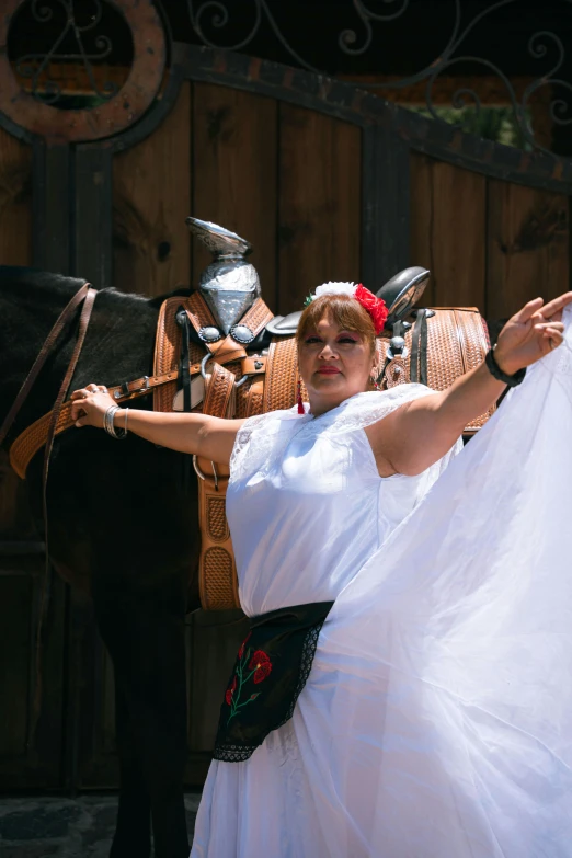 a lady dressed in a frock and traditional garb, is holding a dress with a brown horse nearby