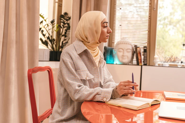 a young muslim woman sits at a table writing and reading a book