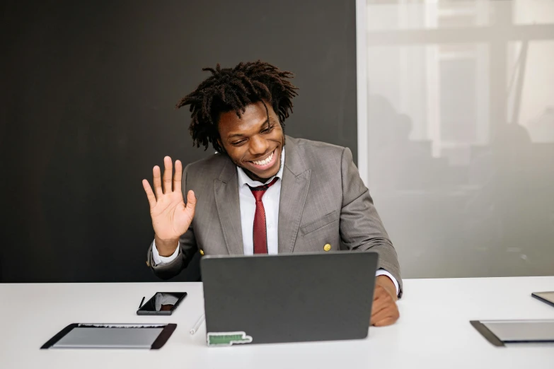 a man dressed in a suit and tie waves to someone behind him in front of a laptop